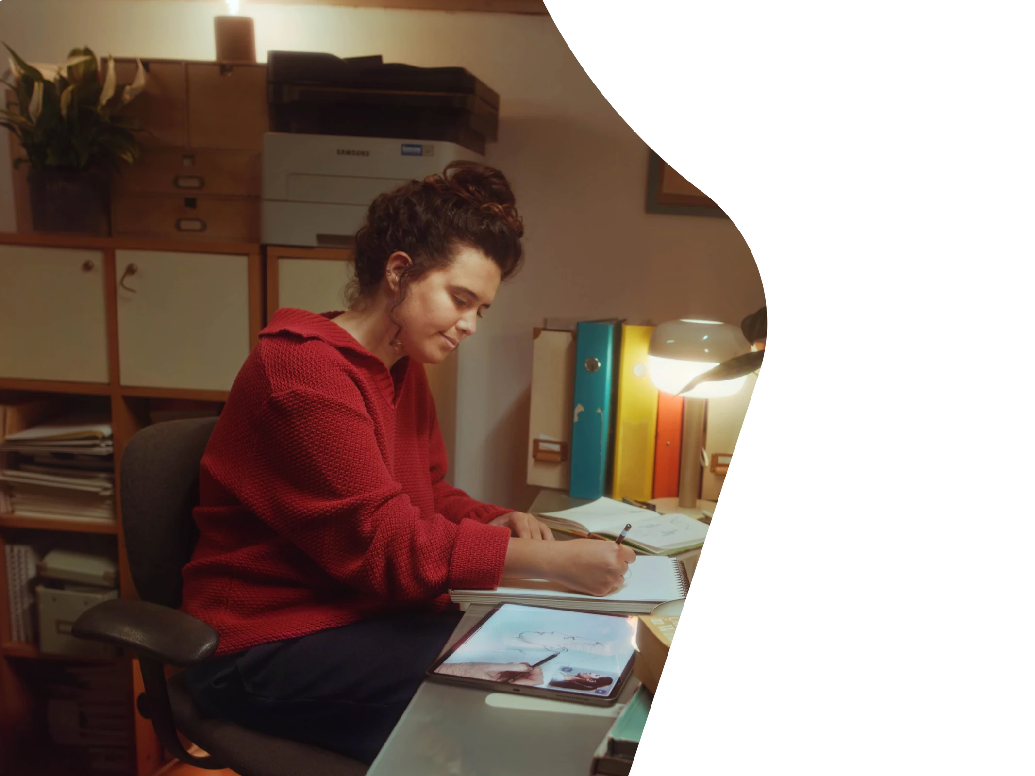 A young woman at a desk in a warm home studio, drawing in a sketchbook with an iPad beside her