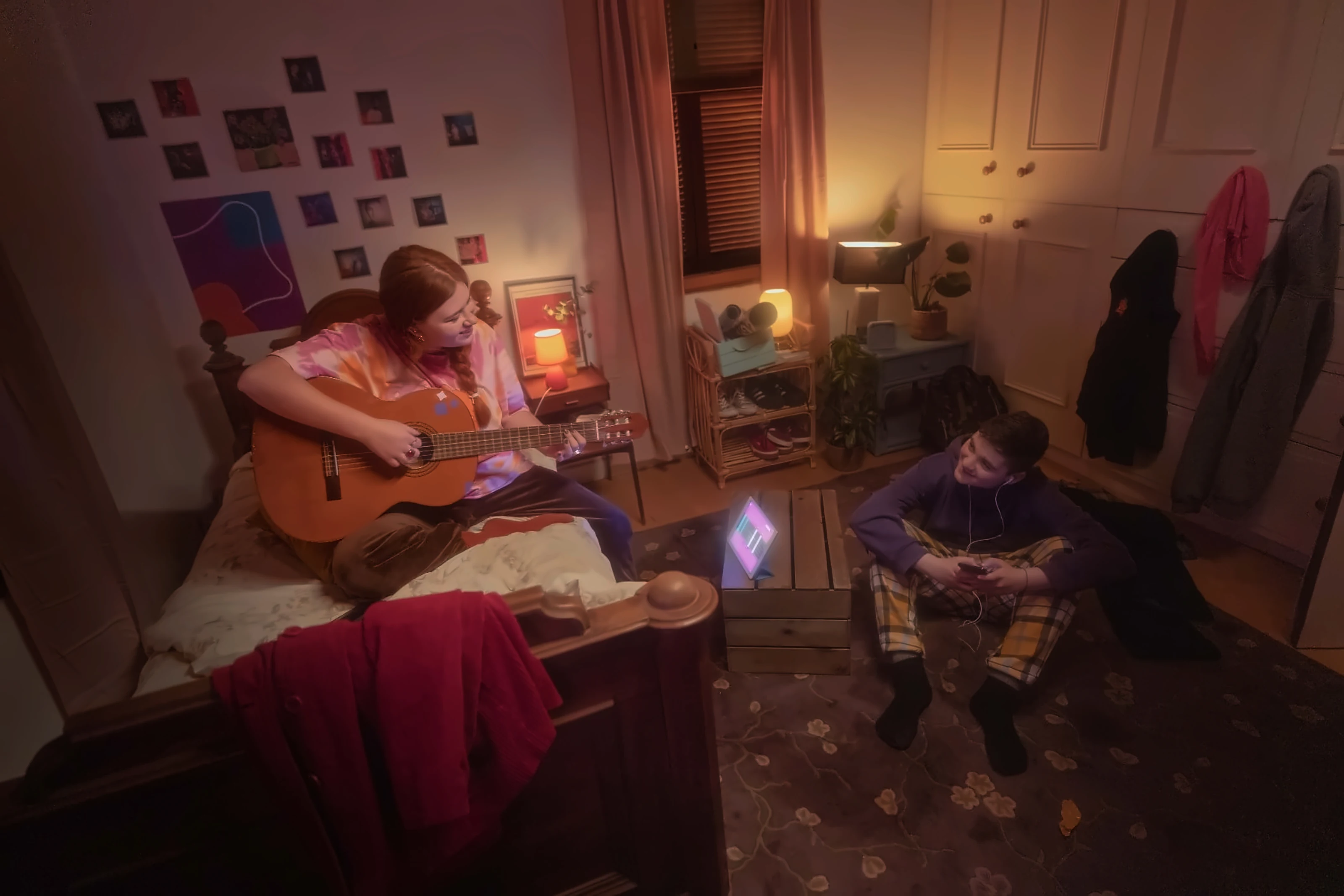 A young woman plays guitar on her bed with Simply Guitar open on a tablet while a friend listens from the floor