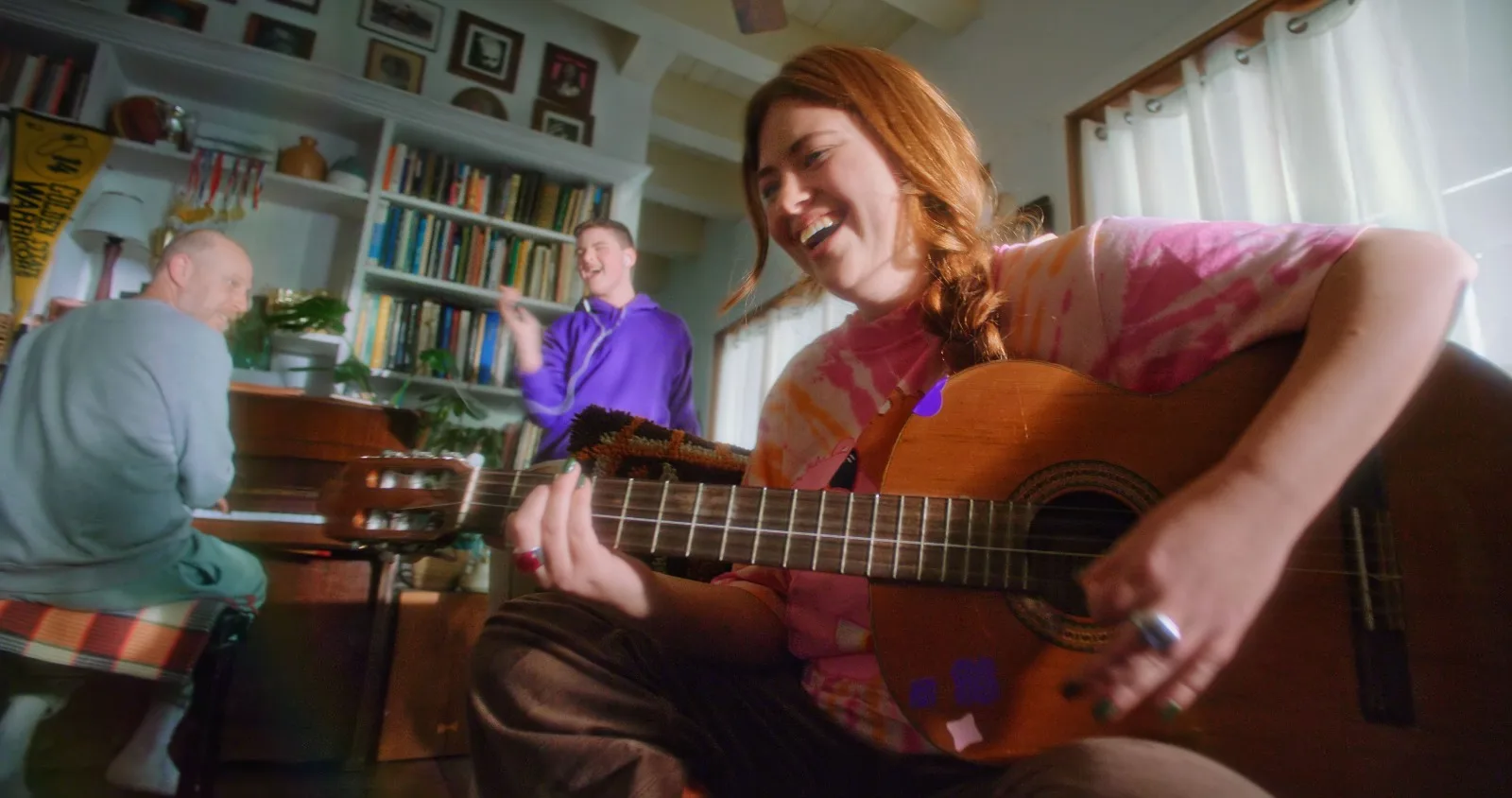 A woman laughs as she plays guitar at home while her family plays piano and sings